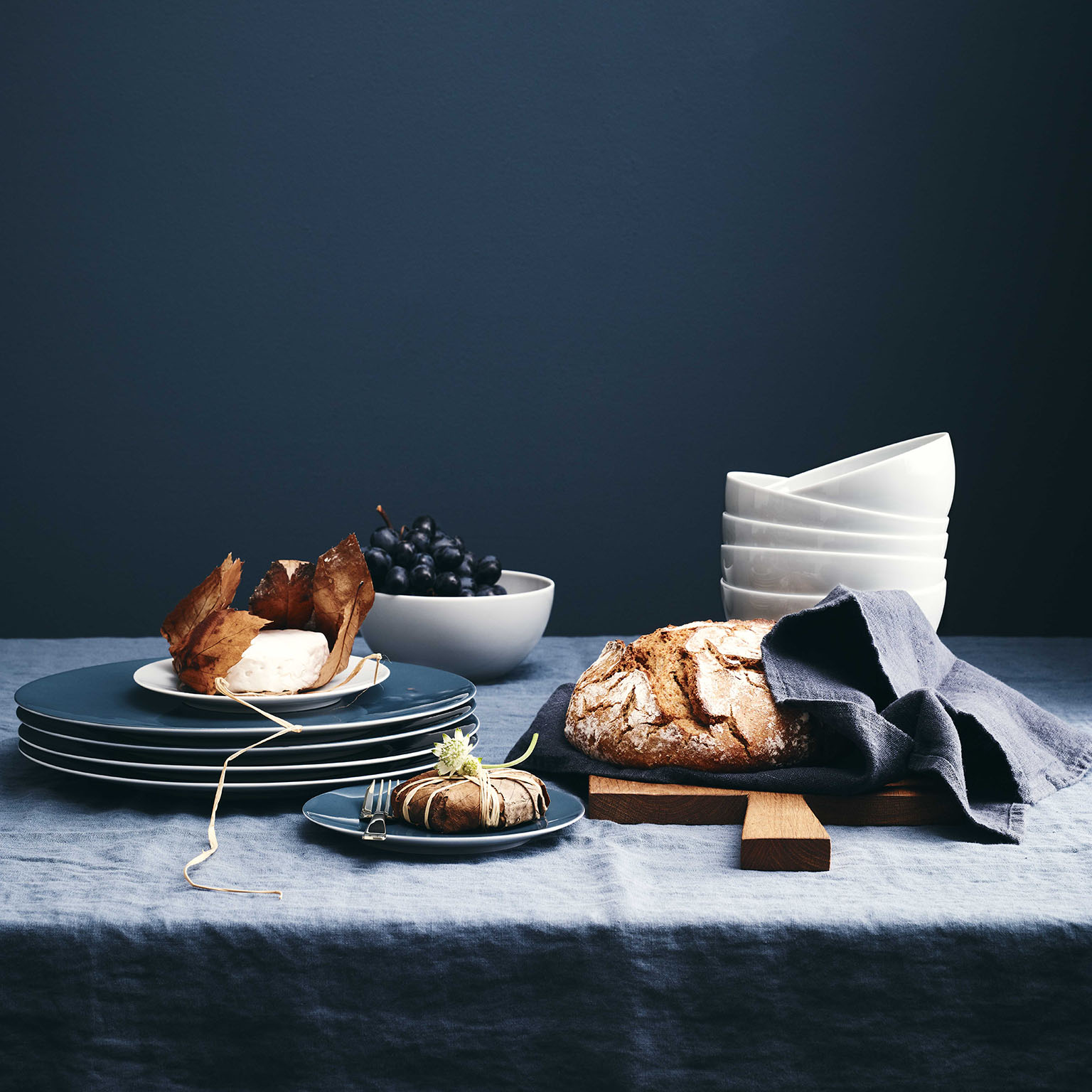 Tisch mit dunkelblauer Leinentischdecke vor dunkelblauem Hintergrund mit Comfort Blue Tellern von TAC Sensual, Weintrauben, Brot und einem Stapel von Schüsseln in TAC Weiss.